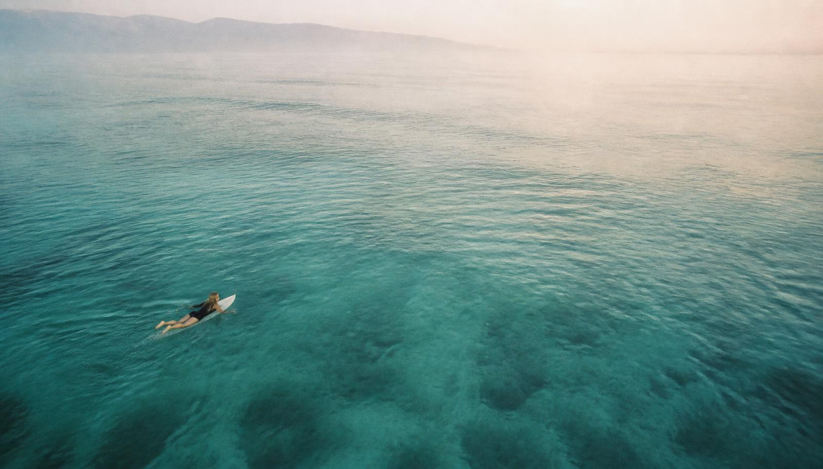 Aerial view of a lone surfer paddling across glassy turquoise water